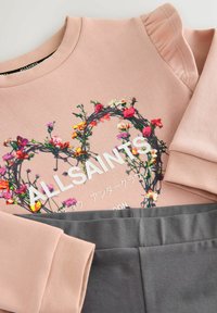 Ružová mikina s kvetinovou výšivkou v tvare srdca, s bielym nápisom a detailom volánovej rukávu, v kombinácii so sivými nohavicami.