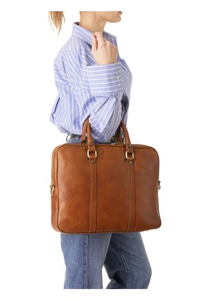 Donna con una camicia a righe blu e jeans tiene una valigetta di pelle marrone per i manici al suo fianco.