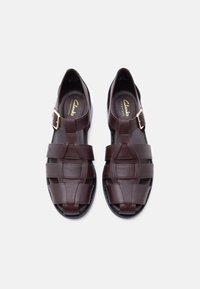 Ζευγάρι καφέ δερμάτινα σανδάλια Clarks με χρυσούς ιμάντες αγκράφας και υφαντό σχέδιο, εμφανιζόμενα από πάνω σε λευκό φόντο.