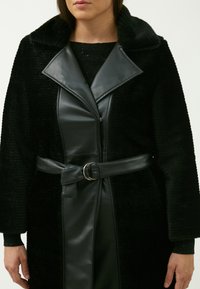Cappotto nero con un esterno in lana texturizzata e rever in pelle liscia; vita cintrata con una cintura in pelle e dettaglio in metallo.