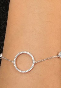 Silverarmband med ett cirkulärt mönster, prytt med små glittrande stenar, sammanlänkade med en delikat kedja. Slät yta.