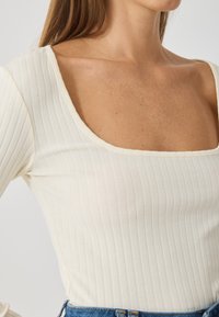 Top cu mânecă lungă, cu model canalat, în alb-off, cu decolteu pătrat. Fabricat dintr-un material elastic, având o siluetă ajustată și o textură netedă.