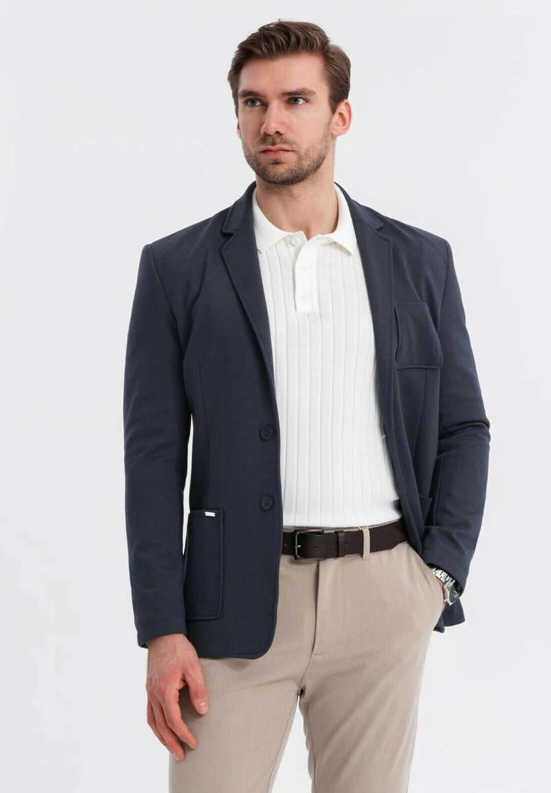 Man i marinblå blazer, vit ribbad polo, beige byxor, brunt bälte och silverfärgad klocka, står med en hand i fickan.