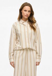 Camisa de botones a rayas beige con acentos amarillos y un bolsillo en el pecho izquierdo, hecha de una tela texturizada; combinada con pantalones sueltos a juego.