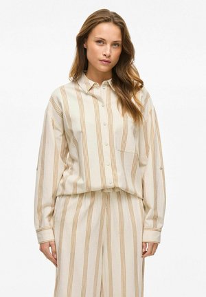 Camisa de botones a rayas beige con acentos amarillos y un bolsillo en el pecho izquierdo, hecha de una tela texturizada; combinada con pantalones sueltos a juego.