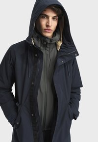 Tamnoplavi parka s kapuljačom, s kragnom obloženim krznom, prednjim kopčanjem na dugmad i džepovima na zip. Nosio se preko sive jakne s visokim ovratnikom.