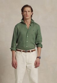 Grønn lin skjorte med rullede ermer, kombinert med hvite jeans og et brunt belte. Klokke på håndleddet, enkel bakgrunn.