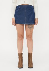 Mørkeblå denim miniskjørt med hvite sømmer, frontlomme og sølvdetaljer. Kombinert med brune ankelstøvler. Glatt tekstur.