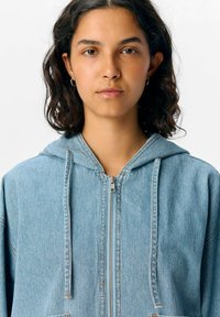 Helles blauer Denim-Zip-Hoodie mit Kapuze und Zugband, Fronttaschen und sichtbaren Nähdetails. Weicher Stoff, lässiger Stil.