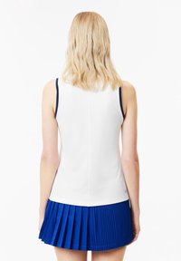 Haut sans manches blanc avec accents bleu marine, tissu texturé. Associe à une jupe plissée bleue, montrant la vue de dos. Finition lisse, design athlétique.