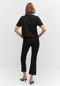 Nainen, jolla on lyhyet vaaleanruskeat hiukset, pukeutuneena mustaan lyhythihaiseen tiukkaan toppiin, mustiin leveälahkeisiin housuihin ja mustiin korkokenkiin, seisoo kädet lanteilla.