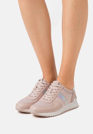 Legs wearing light pink Michael Kors sneakers with iridescent side stripes, white soles, and laces, standing on a white background.
