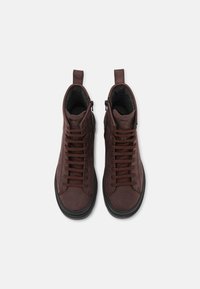 Hnědé kožené vysoké tenisky s plochými tkaničkami, zaoblenou špičkou, polstrovaným límcem a černou gumovou podrážkou. Mají boční zip pro snadné obouvání.