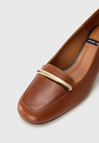 Mocassin en cuir marron avec un bout carré arrondi et un détail de barre métallique dorée brillante sur le cou-de-pied, sur fond gris clair.