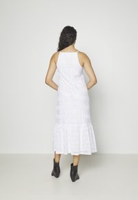 Femme aux longs cheveux foncés portant une robe midi blanche sans manches avec un motif en zigzag et des bottines noires, debout, mains sur les hanches.