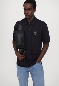 Marineblå polo-shirt med tre-knaps placket og diamanlogo, parret med en sort crossover-taske med lynlås og justerbar rem.