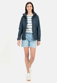 Navyblauwe nylon jas met een capuchon, ritsluiting en twee voorzakken, gedragen over een gestreept shirt en lichte denim shorts. Witte sneakers.