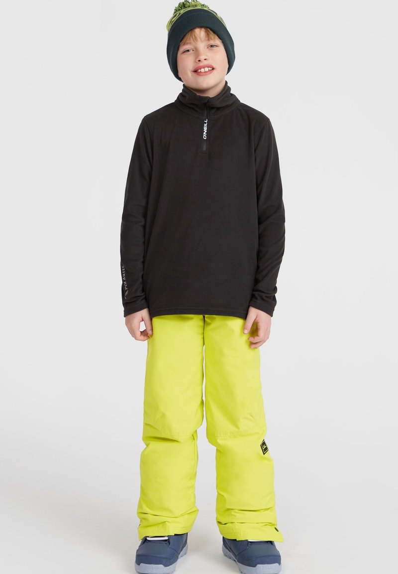 Maglietta nera a maniche lunghe, pantaloni da neve giallo fluorescente, stivali blu e un berretto verde scuro con pompon. Design semplice, abbigliamento outdoor funzionale.