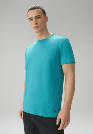 Junger Mann steht und trägt ein einfaches türkisfarbenes kurzärmeliges Hemd und eine schwarze Hose vor einem neutralen grauen Hintergrund.