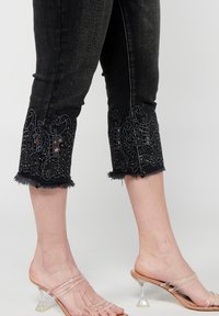 Crne traperice kapri, ukrašene složenim tamnim cvjetnim vezom s perlicama na rubu, sa razmršenim završetkom i prilagođenim krojem.