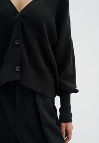 Černý, volný kardigan s výstřihem do V, zapínáním na knoflíky, předními kapsami a žebrovanými manžetami. Vyrobený z měkké, strukturované látky.