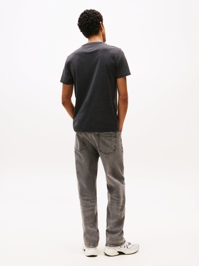 Homme aux cheveux bouclés, de dos, portant un t-shirt gris foncé, un jean gris délavé et des baskets blanches sur fond blanc.