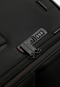 Valigia nera con esterno in nylon testurizzato, doppi zip, serratura a combinazione e dettagli di cucitura rossa per un tocco di stile in più.