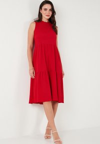 Red sleeveless dress with a high neckline, tiered skirt, and a smooth, textured fabric. Features a knee-length design and relaxed fit.