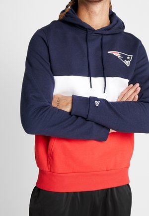 Person iført navyblå, hvide og røde farveblokke hoodie med New England Patriots-logo, krydsede arme og sorte bukser.