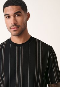 Schwarzes, geripptes T-Shirt mit vertikalen Streifen in Beige und Weiß. Rundhalsausschnitt, kurzärmlig und mit einer strukturierten Stoffoberfläche.