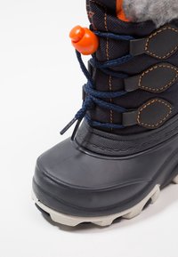 Bota de invierno negra con parte superior de tela texturizada, botón con acento naranja y suela gruesa de goma gris con agarre estampado.