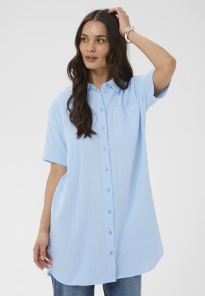 Žena s dlhými tmavými vlasmi, oblečená v svetlomodrej košeli s krátkym rukávom a džínsami, dotýkajúca sa rukou hlavy.