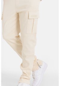 Beige cargobroek met zijzakken, gestructureerd materiaal en ritssluitingdetails bij de manchetten. Draagt met witte sneakers.