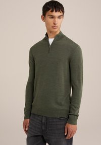 Suéter verde con cremallera en el cuello, hecho de una tela texturizada, con mangas largas y un diseño ajustado, combinado con jeans oscuros.