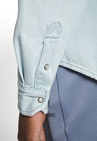 Camicia in denim azzurro chiaro con maniche e polsini con bottoni, caratterizzata da un tessuto testurizzato, orlo grezzo e dettagli in metallo sui bottoni.