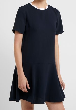 Frau trägt ein kurzärmeliges marineblaues Kleid mit tailliertem Bund und ausgestelltem Rock, zeigt Oberkörper und obere Beine vor einem einfarbigen Hintergrund.