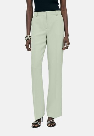 Vrouw met lichtgroene rechte broek, zwarte sandalen met bandjes, zwart mouwloos topje, brede gestreepte armband en juwelenring.