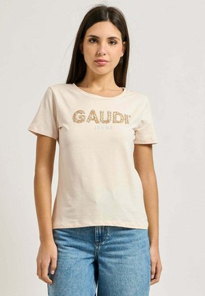 Giovane donna con capelli scuri indossa una maglietta beige a maniche corte con la scritta "GAUDI JEANS" e jeans blu a vita alta, in piedi davanti a uno sfondo neutro.