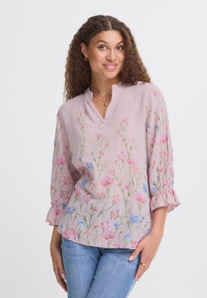 Kvinde med krøllet hår iført en lyserød bluse med blomsterprint og rynkede manchetter samt blå jeans, smilende med hånden på hoften.