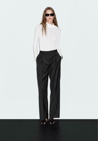 White long-sleeve turtleneck shirt paired with dark gray high-waisted pleated trousers; model wearing black sunglasses and strappy sandals.