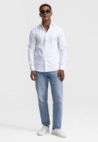 Witte blouse met lange mouwen en knopen, blauwe jeans en witte sneakers; model draagt een donkere zonnebril tegen een effen achtergrond.