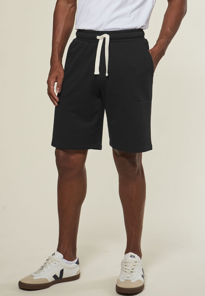 Man wearing black drawstring shorts and white sneakers with black logos, standing against a plain light background.