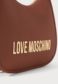 Bolsa de ombro em sintético castanho, com uma textura suave, apresentando o logótipo gold "LOVE MOSCHINO" na frente. Fecho superior com zipper.