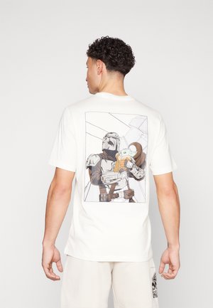 Mann trägt ein weißes T-Shirt mit einer Grafik eines Mandalorianers, der Grogu auf dem Rücken hält, vor einem einfach skizzierten Hintergrund.