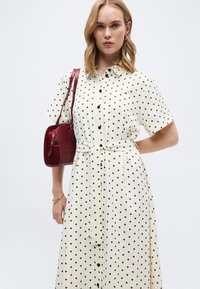 Γυναίκα που φοράει λευκό φόρεμα με κοντά μανίκια και κουμπιά, διακοσμημένο με μαύρα πουά, ζώνη στη μέση, χρυσό βραχιόλι και κρατάει μια σκούρα κόκκινη δερμάτινη τσάντα ώμου.