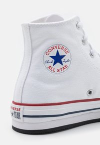Bílé vysoké tenisky z plátna s kulatým logem "Converse All Star" modrou hvězdou. Červené a modré pruhy na gumové podrážce.