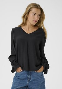 Black long-sleeve blouse with a V-neckline. Loose fit and textured fabric. Slightly gathered sleeves with elastic cuffs. Worn with blue jeans.