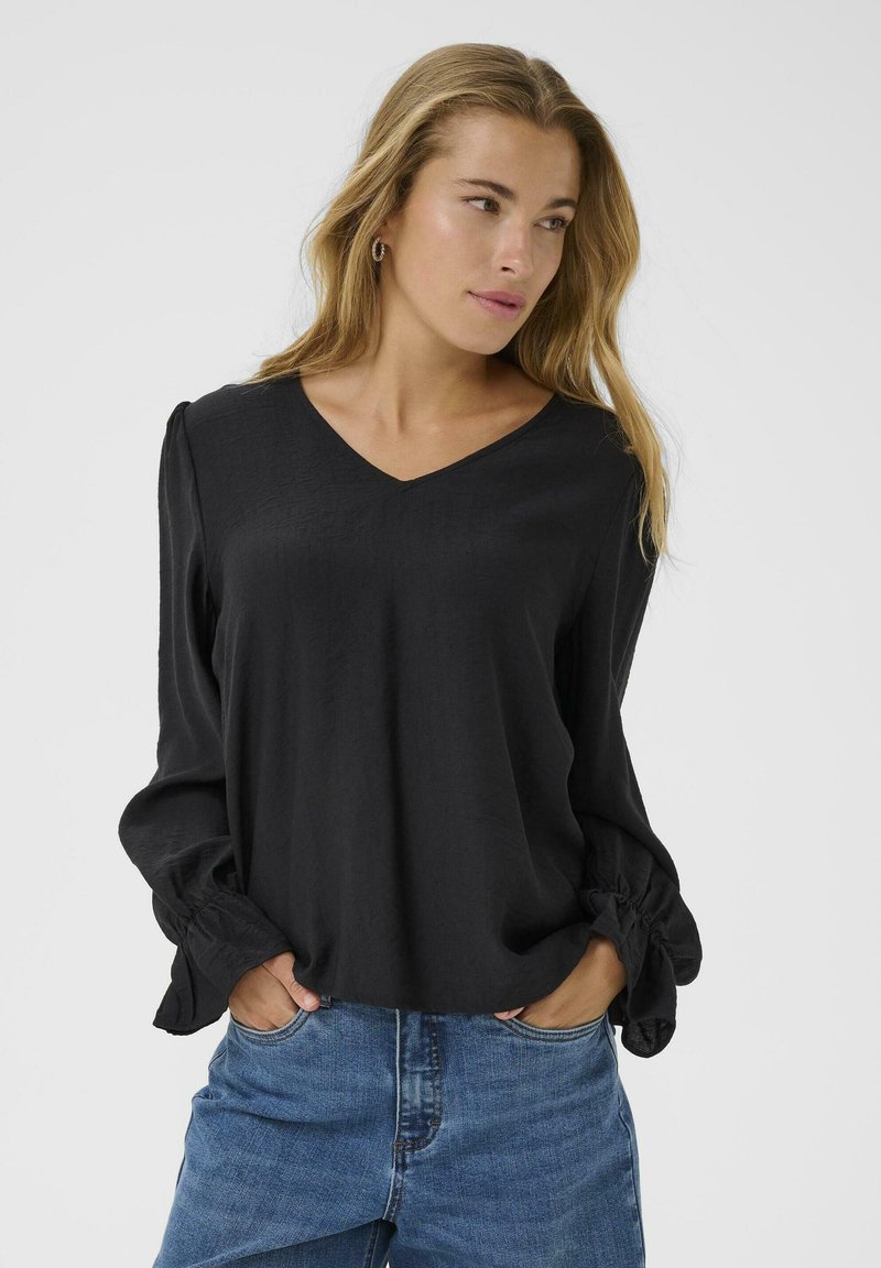 Black long-sleeve blouse with a V-neckline. Loose fit and textured fabric. Slightly gathered sleeves with elastic cuffs. Worn with blue jeans.