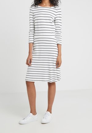 Femme portant une robe blanche au genou avec des rayures horizontales noires et des baskets blanches, debout devant un fond clair uni.