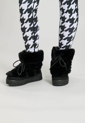 Sorte pelsforede støvler med runde tæer, flade strukturerede såler og sløjfe-accenter på skaftet, båret med sort-hvide houndstooth leggings.